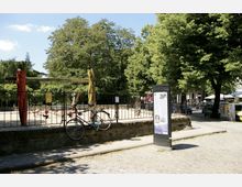 Spielplatz mit Sandfläche, Holzelementen und Schaukeln, umgeben von einem Metallzaun; im Vordergrund ein angelehntes Fahrrad und eine Informationsstele. Im Hintergrund stehen große Bäume, und rechts sind parkende Autos sowie ein Cafébereich sichtbar.