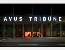 Außenaufnahme der beleuchteten "AVUS Tribüne" bei Nacht, einem Gebäude mit großen weißen Lettern auf der Fassade. Im unteren Bereich sind orange Türen und ein beleuchteter Eingangsbereich zu sehen, vor dem sich Personen aufhalten.