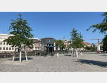 Pflasterplatz mit einer Glas- und Stahl-Konstruktion des Eingangs zur U-Bahn-Station "Museumsinsel", umgeben von jungen Bäumen. Im Hintergrund historische Gebäude mit verzierter Architektur und eine Brücke über einen Kanal.