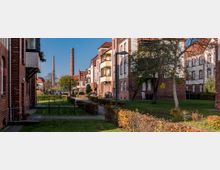Wohnsiedlung mit historischen Backsteingebäuden, umliegenden Grünflächen und Hecken. Im Hintergrund sind zwei hohe Schornsteine zu sehen, die auf eine industrielle Umgebung hinweisen.