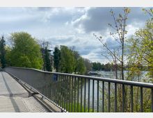 Fußweg auf einer Brücke mit Metallgeländer, das einen Blick auf einen Fluss eröffnet, umgeben von grünen Bäumen und vereinzelten Häusern am Ufer; im Hintergrund sind Boote und ein bewölkter Himmel zu sehen.