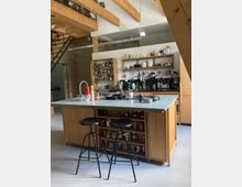 Moderne Wohnküche mit Holzelementen, einer zentralen Kücheninsel mit grüner Arbeitsplatte und integrierten Regalen, bestückt mit Gewürzgläsern. Im Hintergrund befinden sich offene Regale mit Geschirr und Küchenutensilien, freiliegende Holzbalken und eine Treppe im industriellen Stil.