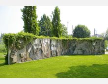 Eine mit Mosaiken verzierte Betonwand in einem parkähnlichen Bereich, überwuchert von grünen Ranken. Im Hintergrund sind Bäume, Sträucher und vereinzelte Gebäude sowie ein sonniger Himmel erkennbar.