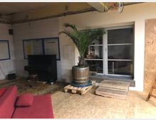 Ein Innenraum mit einem schwarz lackierten Klavier und einem Hocker auf einem Teppich, links an der gefliesten Wand. In der Mitte steht eine große Palme in einem Holzfass, platziert auf einer Holzpalette. Im Hintergrund ist eine Glastür mit Blick in einen angrenzenden Raum erkennbar.