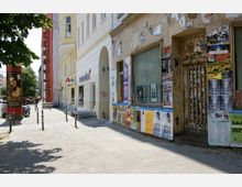 Straßenszene mit einer Mischung aus renovierten und verfallenen Gebäuden; rechts ein Gebäude mit abgenutzter Fassade, beklebt mit Plakaten und Graffiti, links sind modernere gelbe und weiße Häuser sowie ein Apothekenlogo sichtbar. Der Boden besteht aus Kopfsteinpflaster, und im Hintergrund befinden sich Bäume, ein Baugerüst und geparkte Fahrzeuge.