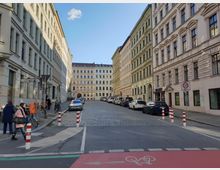 Straßenszene in einem städtischen Wohngebiet mit gepflasterter Fahrbahn und mehrstöckigen, hellen Altbauten auf beiden Seiten. Im Vordergrund ein Fahrradweg, einige Fußgänger sowie abgestellte Autos am Straßenrand.