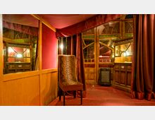 Innenraum eines historischen Theaters oder Cabarets mit Holzverkleidung, roten Vorhängen und Spiegeln. Ein Stuhl mit Leopardenmuster steht auf einem roten Teppich, und im Hintergrund sind geschwungene Fensterrahmen und dezente Lampen sichtbar.