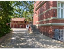 Backsteinbau mit verzierten Details und Treppenaufgang im Vordergrund, daneben ein gepflasterter Weg zu einem Hof mit einem zweistöckigen Gebäude aus Ziegelstein und roten Holztüren. Links befinden sich Bäume und Sträucher, die Schatten spenden.