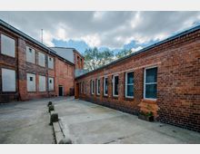 Backsteingebäudekomplex mit einem gepflasterten Innenhof, bestehend aus einem mehrstöckigen Gebäude mit zugemauerten Fenstern und einem einstöckigen Gebäude mit großen Fenstern; im Hintergrund Bäume und ein wolkiger Himmel.