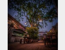 Ein urbaner Außenbereich mit Holzterrasse, Tischen und Stühlen unter einem großen Baum, der mit Lichterketten dekoriert ist. Im Hintergrund sind eine mit Graffiti verzierte Backsteinwand und ein beleuchtetes Gebäude sichtbar.