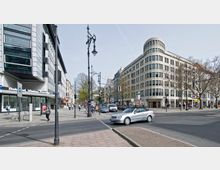 Straßenkreuzung in einer urbanen Umgebung mit modernen Gebäuden; links ein mehrstöckiges Gebäude mit Glas- und Metallfassade, rechts ein abgerundetes Eckgebäude mit heller Steinverkleidung. Im Vordergrund Autos, Fußgänger und mehrere Straßenlaternen, im Hintergrund Bäume und weitere Gebäude.