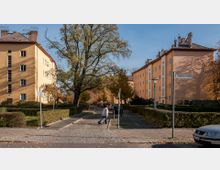 Straßenszene mit mehrstöckigen Wohnhäusern, die in hellgelber Farbe gestrichen sind, und gepflegten Hecken im Vordergrund. Ein großer Baum, ein Straßenschild mit der Aufschrift „Gäblerstraße“ und ein Fußgängerweg mit einer Frau, die einen Einkaufsroller zieht, prägen das Bild.