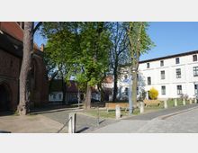 Grüner Platz mit Bäumen und Sitzbänken, umgeben von Backsteingebäuden und einem weißen Wohnhaus. Im Vordergrund sind mit Ketten verbundene Betonpfosten und ein Gehweg aus Kopfsteinpflaster zu sehen.