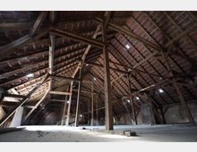 Großer Dachbodenraum mit sichtbarem Holzbalkenwerk und Ziegel-Unterdach, getragen von hohen Stützen und Querstreben. Der Boden ist weitgehend leer, mit gemauerten Außenwänden und mehreren kleinen Dachöffnungen, durch die Licht einfällt.