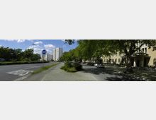 Straßenszene mit breiter Fahrbahn, Busspur und angrenzendem Gehweg; links ist eine Baumreihe mit Grünflächen, rechts stehen beige Wohngebäude mit schattenspendenden Bäumen. Im Hintergrund sind weiße Hochhäuser zu sehen, unter einem blauen Himmel mit vereinzelten Wolken.