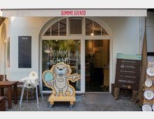 Außenansicht einer kleinen Eisdiele mit dem Namen „Gimme Gelato“. Vor dem Eingang stehen eine Holzfigur eines Tieres mit Eis in der Hand und ein Schild mit Hygieneregeln; daneben befinden sich ein Hochstuhl und Holztische.