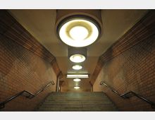 Treppenaufgang in einer U-Bahn-Station mit Wänden aus roten Backsteinen, Metallhandläufen und großen runden Deckenleuchten, die den Bereich beleuchten. Oben ist ein Schild mit der Aufschrift "Ausgang" erkennbar.
