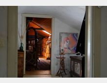 Blick durch eine Tür in ein Dachzimmer mit Holzdecke, Bücherregalen und einem dekorativen asiatischen Standfigur. Im Vordergrund ein Flur mit einem alten Möbelstück, einem stehenden Kleiderständer und einem bunt gemusterten gerahmten Bild an der Wand.