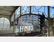 Bahnhof mit überdachter Plattform aus Metall und Glas, versehen mit großen Fenstern und Rundbögen. Im Hintergrund ein modernes Gebäude mit großer Glasfassade, davor Bahngleise und ein Signal mit Aufklebern.