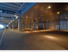 Überdachte Park- oder Ladefläche mit Betonpfeilern, glattem Boden und Wänden aus Mauerwerk. Im Hintergrund sind geparkte Autos, geschlossene Tore und Beleuchtungselemente an der Decke zu sehen.