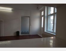 Ein leerer Raum mit weißen Wänden und einem roten Boden, einer großen Fensterfront auf der rechten Seite, durch die Tageslicht einfällt. Im Hintergrund ist eine Türöffnung mit Blick auf einen angrenzenden Raum mit schwarz-weiß kariertem Boden zu sehen, daneben steht ein Papierkorb.