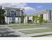 Modernes Regierungsgebäude mit heller Fassade, großen Glasflächen und markanten weißen Säulen. Im Vordergrund befinden sich ein gestreifter Rasen- und Gehwegbereich sowie eine Metallskulptur, umgeben von Bäumen.