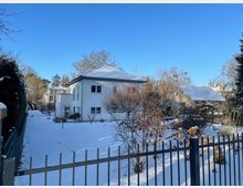 Schneebedeckter Garten mit Sträuchern und kahlen Bäumen vor einem weißen Wohnhaus mit Walmdach und mehreren Fenstern. Im Vordergrund verläuft ein Metallzaun, im Hintergrund sind weitere Häuser und Bäume unter klarem blauem Himmel zu sehen.