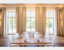 Ein heller, elegant eingerichteter Raum mit großen Fenstern, durch die Tageslicht einfällt, und beigen Vorhängen. In der Mitte steht ein gedeckter Holztisch mit weißen Stühlen, Geschirr, Gläsern und Wasserflaschen.