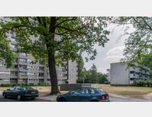 Wohnsiedlung mit mehrstöckigen Wohngebäuden im Stil der Nachkriegsmoderne, umgeben von Rasenflächen und Bäumen. Im Vordergrund parken Autos am Straßenrand, während eine Person auf einer freien Grünfläche sichtbar ist.