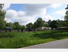 Ländliche Szene mit grüner Wiese im Vordergrund, einem eingezäunten Areal mit Holztoren und einer kleinen, flachen Betongebäude im Hintergrund. Umgeben von Bäumen und bewölktem Himmel.