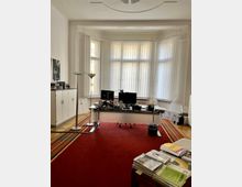 Helles Büro mit einem großen Schreibtisch, zwei Monitoren und Büromaterialien vor einer Fensterfront mit weißen Lamellenjalousien. Der Raum hat hohe Decken, dekorative Stuckelemente sowie einen roten Teppichboden und weiße Wände.