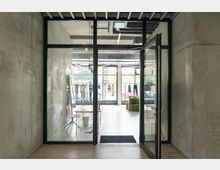 Innenansicht eines modernen Showrooms mit Betonwänden, einer Glastür und großen Fenstern. Im Raum stehen Designermöbel, und Kleidung auf Kleiderständern ist ausgestellt.