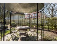 Garten mit einer kleinen Sitzgruppe aus Metall unter einer offenen, kuppelförmigen Pergola. Im Hintergrund sind eine Hecke, Bäume und Wohnhäuser mit roten Ziegeldächern zu sehen.