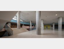 Weitläufiges, modernes Innenraumdesign mit hellem Bodenbelag, großen Betonpfeilern und einer offenen Treppenkonstruktion. Die Decke ist schlicht und mit eingelassenen Leuchten versehen, während die umlaufenden Wände Fensterfronten und farbige Akzente wie gelbe und blaue Flächen aufweisen.