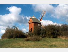 Eine historische Windmühle aus Holz mit roten Wänden und weißen Flügeln steht inmitten einer Wiesenlandschaft, umgeben von Büschen und vereinzelten Bäumen unter einem wolkigen Himmel.