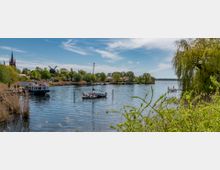 Flusslandschaft mit ruhigem Wasser, umrahmt von Bäumen und Schilf. Links im Hintergrund sind eine Kirche mit spitzem Turm, eine Windmühle und einige Häuser zu sehen; Boote liegen am Ufer und auf dem Wasser vor Anker.