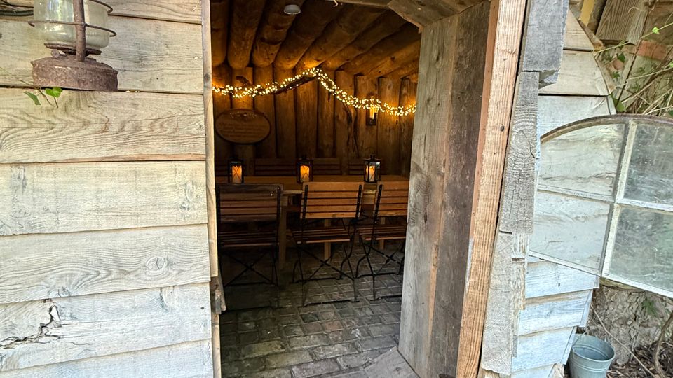 Blick durch eine offene Holztür in eine kleine, rustikale Holzhütte mit Tonnenform, Holzbalkendecke und Steinboden. Im Inneren stehen Holzklappstühle, dekorative Laternen und eine Lichterkette; außen sind Holzverschalung, Efeu und ein altes Lampengehäuse zu sehen.