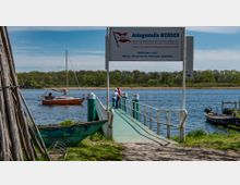 Ein Anlegesteg in Werder an einem Fluss oder See, mit einem Schild mit Informationen zu Abfahrten. Im Hintergrund ist ein kleines Segelboot auf dem Wasser und eine bewaldete Uferzone sichtbar, während im Vordergrund Holzpfähle und ein altes Boot am Ufer stehen.