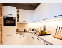 Moderne Küche mit weißen Einbauschränken, einer integrierten Mikrowelle und einem Kochfeld. Die Arbeitsfläche zeigt Elektrogeräte wie Wasserkocher und Kaffeemühle, daneben ein Schneidebrett mit Obst und ein Messerblock. Im Hintergrund sind Ziegelwände und eine indirekte Beleuchtung zu sehen.