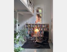 Gemütlicher Innenraum mit Bücherregal, einer Stehleuchte und modernen Sitzmöbeln aus schwarzem Leder. Im Hintergrund ein Fenster mit Rundbogen, das den Blick auf ein Backsteingebäude draußen freigibt, sowie Pflanzen im Vordergrund und ein offener Galeriebereich oben.