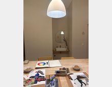 Moderner Innenraum mit langem Holztisch, darauf Zeichen- und Bastelmaterialien sowie Schalen; darüber hängt eine große weiße Pendelleuchte. Im Hintergrund ein kleines Waschbecken mit rundem Spiegel und ein Türbereich mit Toilettenpiktogramm an einer hellen Wand.