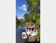 Ein Hausboot mit einer begrünten Terrasse liegt an einem ruhigen Flussufer, umgeben von üppigem Grün. Daneben befindet sich ein kleines Ruderboot im Wasser, und im Hintergrund sind zeitgenössische Gebäude sichtbar.
