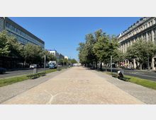 Breiter Fußgängerweg zwischen zwei von Bäumen gesäumten Straßen in einer städtischen Umgebung, flankiert von modernen und klassischen Gebäuden. Entlang des Weges stehen vereinzelt Bänke, und es sind Autos sowie ein LKW auf den Straßen zu sehen.