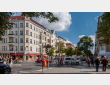 Straßenszene an einer Kreuzung mit Mehrfamilienhäusern im Altbaustil, Geschäften und Cafés im Erdgeschoss. Im Vordergrund sind Passanten, Fahrradständer und Fahrzeuge zu sehen, umgeben von Bäumen bei sonnigem Wetter.