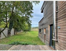 Holzhaus mit vertikaler Holzverkleidung und einer großen Glas-Schiebetür, daneben ein glänzendes Metallrohr. Im Vordergrund eine Holzterrasse, im Hintergrund ein Garten mit grünem Rasen, Bäumen und einem Holzzaun.