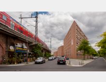 Straßenszene mit einer erhöhten Bahntrasse aus Backstein, auf der ein roter Regionalzug fährt, einem Fischrestaurant unter der Trasse und einem großen, kantigen Backsteingebäude rechts. Geparkte Autos säumen die Straße, und Bäume stehen entlang des Gehwegs.