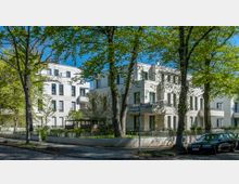 Mehrfamilienhäuser mit heller, moderner Fassade in einer ruhigen Wohngegend, umgeben von hohen Bäumen und eingerahmt von einem Zaun. Im Vordergrund verläuft eine gepflasterte Straße mit parkenden Autos.