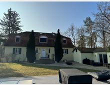 Gelbes zweistöckiges Haus mit rotem Walmdach, Dachfenstern und grünen Fensterläden, umgeben von hohen Bäumen. Im Vordergrund eine Pflastersteintreppe, ein kleiner Garten und eine Terrasse mit abgedeckten Möbeln.