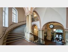 Treppenhaus eines historischen Gebäudes mit geschwungener Steintreppe, verzierten Balustraden und Säulen. Im Hintergrund befinden sich Rundbögen, eine große Uhr und ein Eingangsbereich mit verglasten Doppeltüren.