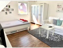 Helles Wohnzimmer mit Holzfußboden, einem Einzelbett auf der linken Seite, einer weißen Couch mit Kissen, einem kleinen Couchtisch und einem grauen Teppich. Im Hintergrund steht ein weißer Kleiderschrank mit Glastüren, und durch ein Fenster fällt Tageslicht ein.